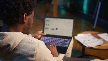 African american freelancer working from home using financial graphs, reviewing statistical data to compare supply and demand numbers. using business charts and writing on notepad. Camera B. video