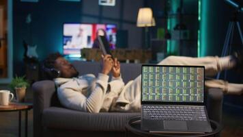 African american person using his tablet laying on the couch while he pays attention to laptop with stock market data trends. Focused man comparing trading graphs numbers before investing. Camera B. video