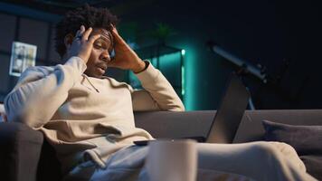 Black person having health problems suffering from headache and nausea, sitting in the living room and struggling to work on tasks. Remote worker suffering from illness at home. Camera B. video
