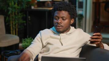 Black freelancer does his research by reading articles and data driven papers at home, using phone and pc to collect information for a new project. Man multitasking and studying files. Camera A. video
