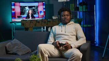Cheerful man in his apartment playing games on couch, enjoying day off from work. Black gamer battling enemies in online multiplayer shooter on gaming console. Camera A. video