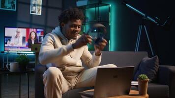 African american male person listening to a podcast and multitasking on the coffee table, browsing the internet and navigating through his laptop with headphones on. Self employed man. Camera B. video