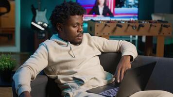 Young adult working remotely from his loft apartment on a laptop. Black freelancer focusing on tasks, blending productivity and relaxation while browsing the web from a cozy sofa. Camera B. video