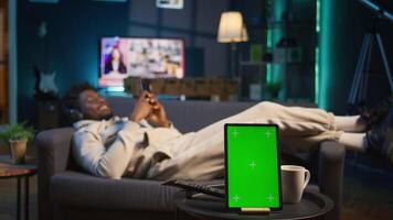 African american young adult laying on the sofa next to mockup copy space, relaxing and browsing web on his smartphone. Happy relaxed person on the couch feels happy with remote job. Camera B. video