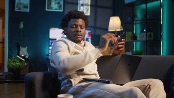 Black freelancer does his research by reading articles and data driven papers at home, using phone and pc to collect information for a new project. Man multitasking and studying files. Camera B. video