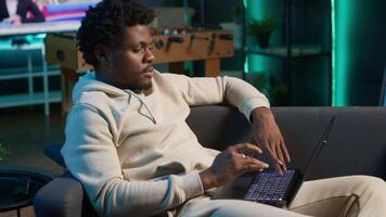 Positive african american man multitasking on a laptop in stylish loft, fully connected to the internet while managing his freelance work and remote responsibilities. Browsing web at home. Camera A. video