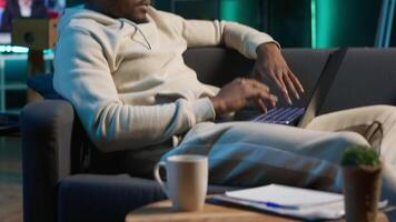 African american person navigating the internet on laptop at his modern luxury loft, relaxing on the couch and browsing web. Cheerful man does freelance work from home. Camera A. video