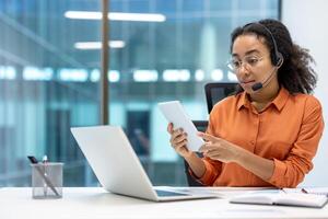 Serious woman with headset and tablet computer working inside office at workplace sitting. Online support worker consulting and helping clients remotely. photo