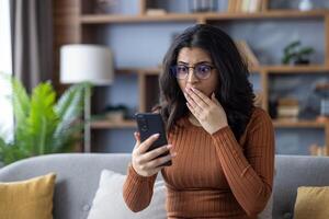Close-up photo of a sad young Indian woman sitting on the sofa at home, looking at the phone in shock, covering her mouth with her hand.