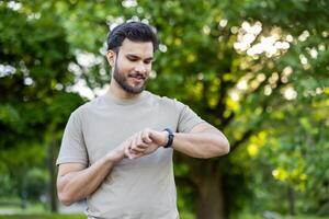 Man engaged in active physical exercises in park, checking fitness tracker with a smile. Surrounded by lush green trees and sunshine, showcasing motivation and health. photo