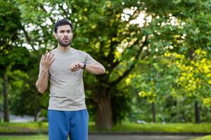 Young man standing in park checking smartwatch expressing dissatisfaction with workout results. Outdoor with green trees in background conveys concept of fitness, effort, and exercise frustration. photo