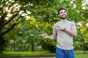 Happy man with smartwatch pointing in green park setting, sunlight filtering through trees. Relaxed and casual, exuding joyful energy and positivity outdoors. Embraces simplicity and connection photo