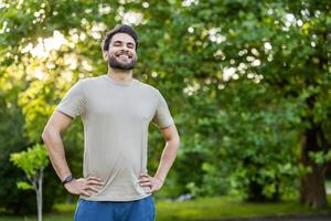 Photo of a smiling young Indian man standing in the park with his eyes closed, hands on his waist and resting after exercising.
