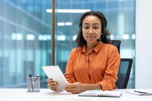 Portrait of online support worker. Woman with headset and tablet computer looking at camera, working inside office at workplace. photo