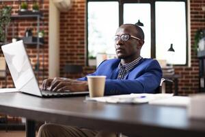 African american male entrepreneur using his laptop and reviewing business plans at office desk. Black businessman in wheelchair researching strategy ideas on digital device in brick wall room. photo