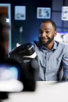 Smiling african american man presenting VR goggles to audience, filming with professional camera. Content creator trying virtual reality headset product before recommending it to fans photo