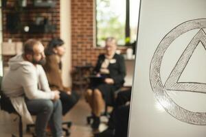 Closeup of aa meeting symbol on white board with ongoing therapy session in background. People engaging in discussions about alcohol addiction with guidance from retired therapist in brick wall office photo