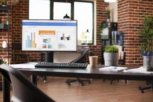 Vacant office desk holds computer displaying company research data on screen. Empty brick wall workspace, arranged with indoor plants, files, and desktop pc on table, waiting to be used by employees. photo