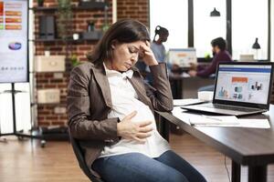 Tired expectant mother with hand on forehead, taking break and looking at her pregnancy belly in startup office. Caucasian pregnant woman sitting at desk, exhausted from researching data on laptop. photo