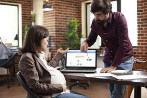 Bearded man pointing out error in company analysis on laptop, during meeting with pregnant white woman. Male and female coworkers using digital device to discuss financial strategy of startup office. photo