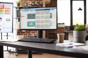 Empty workspace with desktop pc and big digital screen desk displaying business analytics data. Office table with nobody, has computer screen showing company statistics information in brick wall room. photo