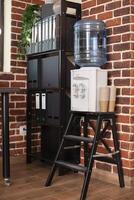Modern workspace containing water cooler for refreshments and cabinet for folders. Brick wall office corner organized with shelves and dispenser, thus representing clean and tidy work area. photo
