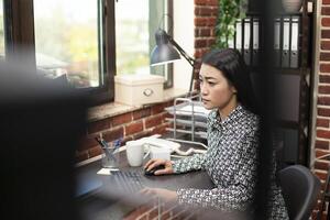Female researcher near window types diligently, updating marketing analytics in brick wall office. Asian woman focuses on project updates at her desk, working on her computer in a modern workspace. photo