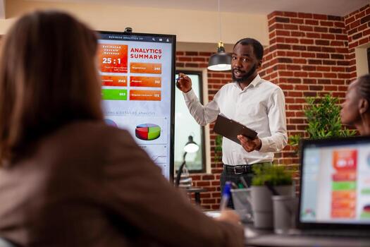 Businessman with clipboard in hand, using digital monitor to explain analytics summary in meeting. African american entrepreneur doing project presentation, planning marketing strategy with his team. photo