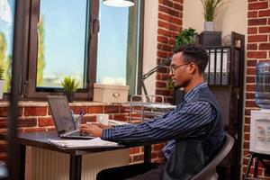 Young businessman sitting at desk and using his laptop for company data research. African american male entrepreneur working from home, gathering startup information on his personal computer. photo