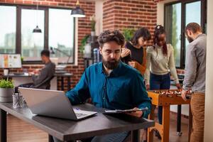 Male office worker focused on reviewing documents and charts on clipboard at desk, laptop placed beside him in brick wall workspace. Young man sitting at table and reading company guidelines. photo