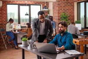 Caucasian man with laptop reviews online documents with african american coworker at table in modern workspace. Male colleagues remain focused on achieving business objectives using digital device. photo