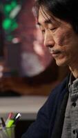Vertical Senior programmer in AI startup office putting on headphones, refining machine learning models to enhance automation. Asian CTO manager listening music while working, camera B close up video