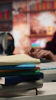 Vertical Focus on stack of books on startup office desk with woman in blurred background typing on PC, solving tasks. Pile of books in venture company workspace in front of employee working, camera A closeup video