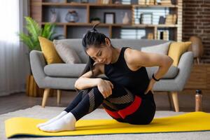 Asian woman sitting on yoga mat at home, holding lower back and showing expression of pain. Indoor scene highlights concepts of back injury, fitness, and wellness challenges. photo