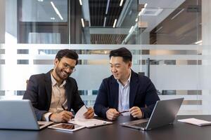 Two professionals are working together, reviewing documents in a modern conference room setting. They are surrounded by laptops and other tools, engaging in a discussion. photo