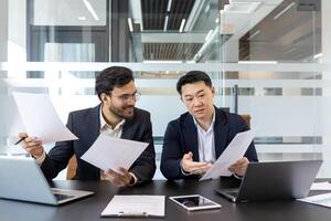 Two business professionals engaging in discussion while reviewing important documents. Workspace includes laptops, tablet, and papers. Office setting promotes teamwork, collaboration photo