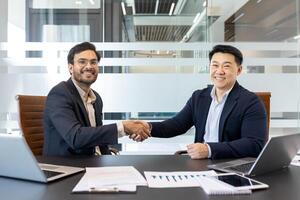 Handshake, two businessmen inside the office, business meeting agreeing on a contract, men smiling happily and looking at the camera, working with a laptop in a meeting room inside the office. photo