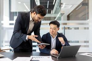 Two businessmen discussing a work project, a man in a business suit at his workplace inside the office. An employee demonstrates a work program on a tablet computer. photo