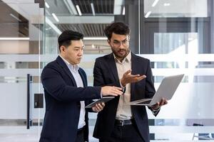 Two businessmen engaged in discussion holding laptop and tablet in modern office. Displaying teamwork, communication in professional business environment. Emphasis on collaboration, technology photo