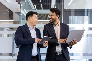 Confident business professionals engaged in friendly conversation using tablet and laptop. Image captures modern workplace, teamwork, and professional attire, highlighting collaboration photo