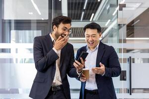 Two colleagues sharing a light moment in office while watching a funny on smartphone. Both men dressed in business suits, one holding a coffee cup, expressing laughter and surprise. photo