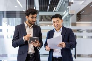 Two business professionals in office discussing reports. One holds documents while the other uses a tablet. Scene depicts collaboration, communication, and teamwork in a professional setting. photo