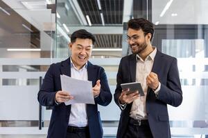 Two business professionals sharing moment of success in office. One holding paperwork, other with tablet. Expressions of joy and achievement visible on faces. Teamwork and collaboration photo