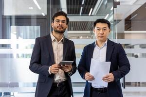 Two business professionals standing in modern office holding documents and tablet. Serious expressions indicate focus on business project. Office setting suggests teamwork and professionalism. photo