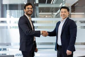 Two businessmen in suits exchanging a confident handshake in modern office. They are smiling, indicating successful collaboration or partnership. Image captures concepts of business success photo