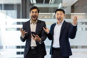 Two businessmen in formal attire expressing excitement and surprise in modern office environment. One holds smartphone, emphasizing technology and achievement. Perfect representation of teamwork photo