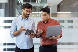 Two diverse office workers discussing work project, serious men in casual clothes standing with tablet computer and laptop, testing new software. photo