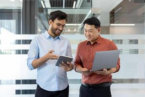 Two programmer colleagues in casual clothes discussing a new software project, men standing by the window with a laptop, smiling, talking. photo