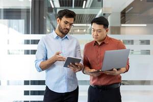 Two male office workers collaborate in modern office, using digital devices. Team discussion, sharing ideas, and analyzing data on laptop and tablet. Professional interaction business environment. photo