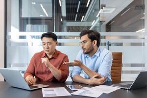 Office workers discussing work project looking at laptop screen, two diverse men sitting at desk with computer, serious and focused in teamwork. photo